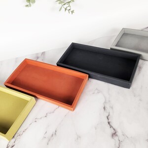 May include: Four rectangular trays in different colors: yellow, orange, black, and gray. The trays are arranged in a row on a white marble surface.