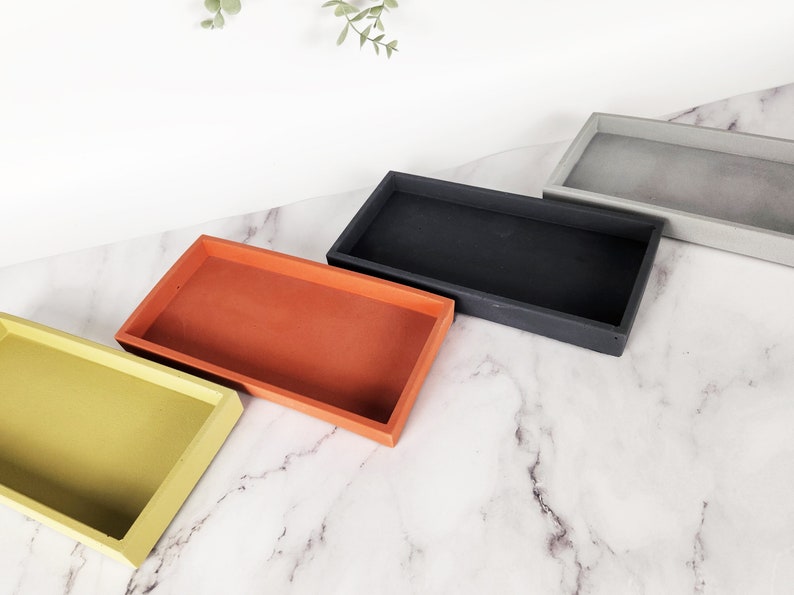 May include: Four rectangular trays in different colors: yellow, orange, black, and gray. The trays are arranged in a row on a white marble surface.