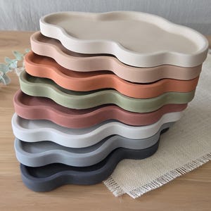 May include: A stack of cloud-shaped trays in various colors, including beige, pink, orange, green, and gray. The trays are made of a solid material and have a wavy edge design. They are stacked on a wooden surface.
