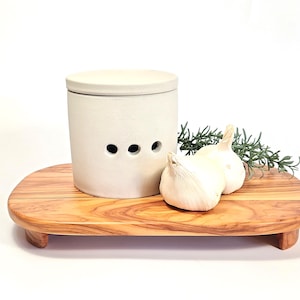 May include: A white ceramic garlic keeper with a lid and three air holes sits on a wooden chopping board with two whole garlic bulbs and a sprig of rosemary.