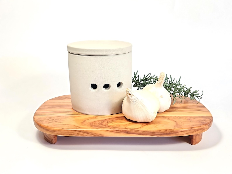 May include: A white ceramic garlic keeper with a lid and three air holes sits on a wooden chopping board with two whole garlic bulbs and a sprig of rosemary.