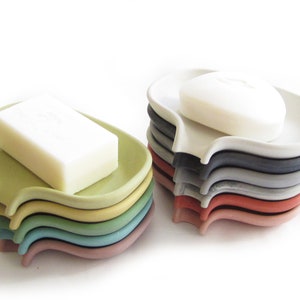 May include: A stack of colorful soap dishes in various shades of green, blue, red, and gray. The dishes are made of a smooth, hard plastic and have a curved shape. A bar of white soap is sitting on top of one of the dishes.