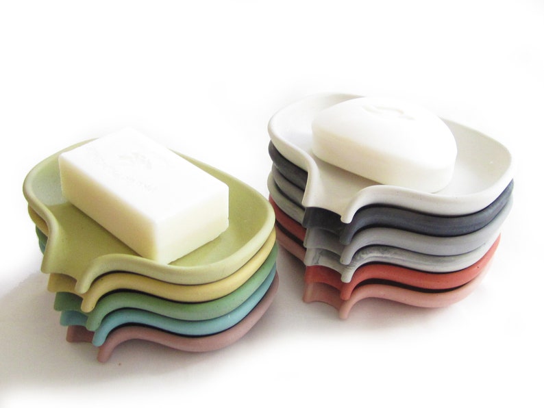May include: A stack of colorful soap dishes in various shades of green, blue, red, and gray. The dishes are made of a smooth, hard plastic and have a curved shape. A bar of white soap is sitting on top of one of the dishes.