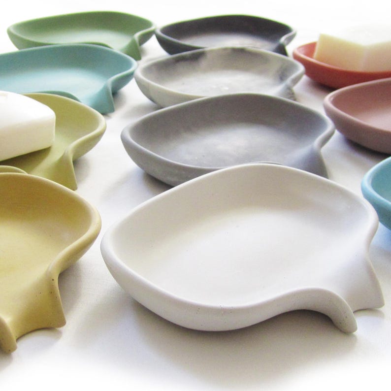 May include: A set of 10 colorful soap dishes made of concrete. The dishes are shaped like a leaf and have a slight curve to them. They are all different colors, including green, blue, gray, white, and yellow.