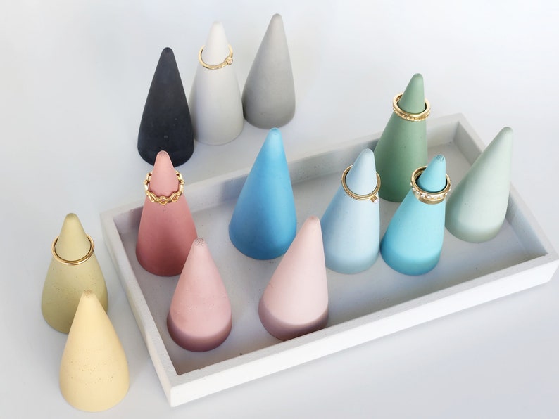 Pu&ograve; includere: Una collezione di porta anelli a forma di cono in vari colori opachi tra cui giallo, rosa, blu, verde, grigio e nero, disposti su un vassoio rettangolare bianco. Diversi anelli in metallo dorato sono esposti sulle punte dei coni.