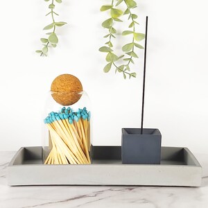 May include: A gray concrete tray with a glass jar filled with matches and a gray concrete incense holder. The jar has a cork stopper and the matches have blue tips. The incense holder has a single incense stick.