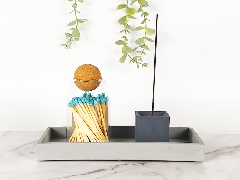 May include: A gray concrete tray with a glass jar filled with matches and a gray concrete incense holder. The jar has a cork stopper and the matches have blue tips. The incense holder has a single incense stick.