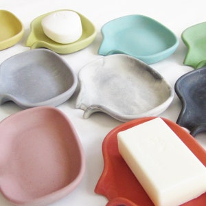 May include: Assortment of ten small, ceramic soap dishes in various colors, including gray, green, blue, yellow, red, and white. The dishes are shaped like a fish with a curved tail and a small indentation in the center to hold a bar of soap.