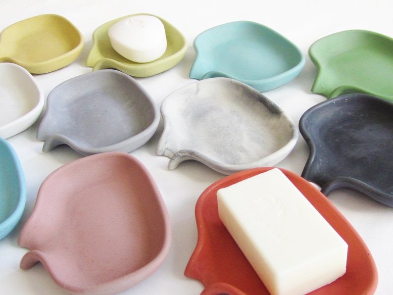 May include: Assortment of ten small, ceramic soap dishes in various colors, including gray, green, blue, yellow, red, and white. The dishes are shaped like a fish with a curved tail and a small indentation in the center to hold a bar of soap.