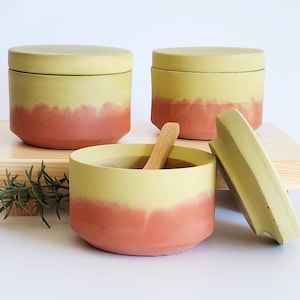 May include: Three-piece ceramic container set with a yellow and coral ombre design. The set includes two lidded jars and one open bowl with a wooden spoon. The lids and bowls have a rounded shape. The set is displayed on a wooden surface.