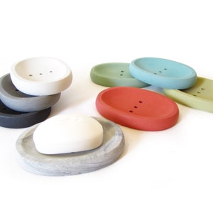 Puede incluir: Un juego de seis jaboneras ovaladas en varios colores, incluyendo gris, blanco, verde, azul y rojo. Las jaboneras est&aacute;n hechas de hormig&oacute;n y tienen un acabado liso y redondeado.