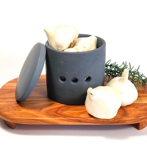 May include: Grey ceramic garlic keeper with a lid, filled with garlic bulbs. The keeper has three holes for ventilation. The keeper is sitting on a wooden serving tray.