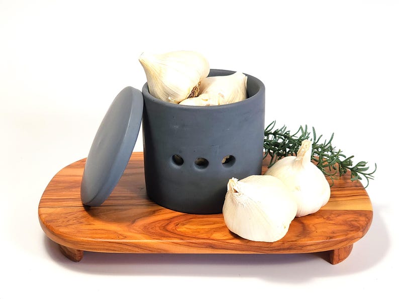 May include: Grey ceramic garlic keeper with a lid, filled with garlic bulbs. The keeper has three holes for ventilation. The keeper is sitting on a wooden serving tray.