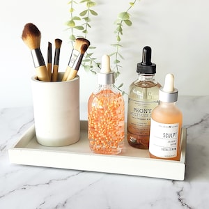 May include: A white tray with a white concrete holder with makeup brushes, a clear glass bottle with orange beads, a clear glass bottle with a black dropper labeled "PEONY MULTI-USE OIL", and a clear glass bottle with a silver dropper labeled "SCULPT COLLAGEN FACIAL SERUM".