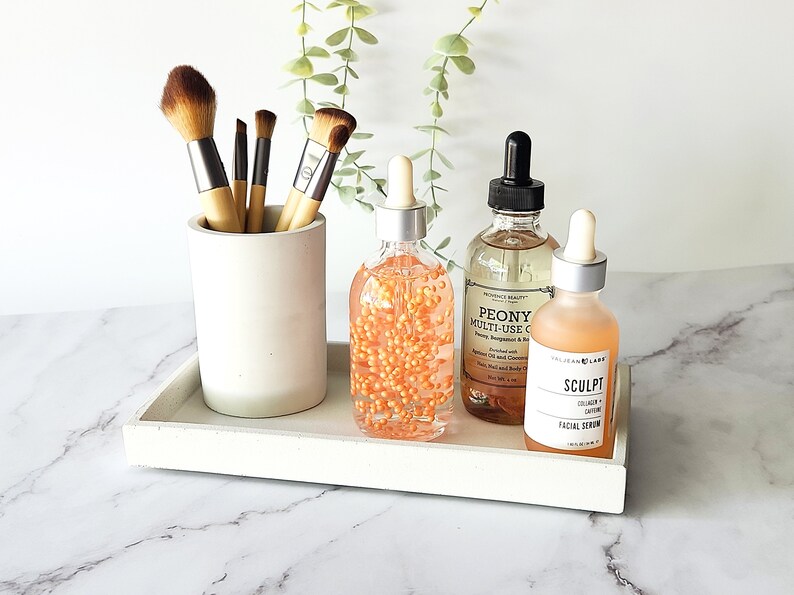 May include: A white tray with a white concrete holder with makeup brushes, a clear glass bottle with orange beads, a clear glass bottle with a black dropper labeled "PEONY MULTI-USE OIL", and a clear glass bottle with a silver dropper labeled "SCULPT COLLAGEN FACIAL SERUM".