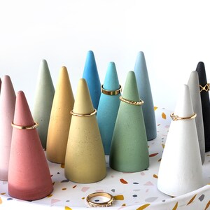 Pu&ograve; includere: Una collezione di dodici porta anelli a forma di cono in vari colori opachi tra cui rosa, giallo, blu, verde, bianco, grigio e nero. Diversi anelli in metallo dorato sono esposti sui coni e un anello poggia sul vassoio in terrazzo.