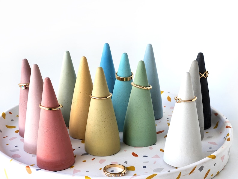 Pu&ograve; includere: Una collezione di dodici porta anelli a forma di cono in vari colori opachi tra cui rosa, giallo, blu, verde, bianco, grigio e nero. Diversi anelli in metallo dorato sono esposti sui coni e un anello poggia sul vassoio in terrazzo.