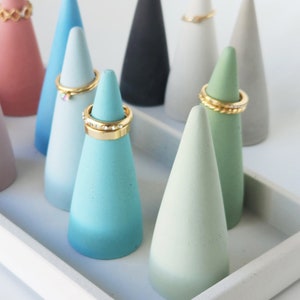 Pu&ograve; includere: Una collezione di coni portaanelli in cemento multicolore nelle tonalit&agrave; rosa, blu, verde, nero e grigio, disposti su un vassoio rettangolare in cemento. Diversi coni sono sormontati da anelli in metallo dorato con vari design.