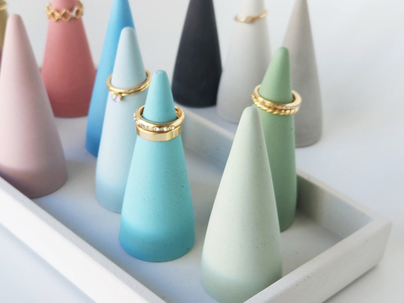 Pu&ograve; includere: Una collezione di coni portaanelli in cemento multicolore nelle tonalit&agrave; rosa, blu, verde, nero e grigio, disposti su un vassoio rettangolare in cemento. Diversi coni sono sormontati da anelli in metallo dorato con vari design.