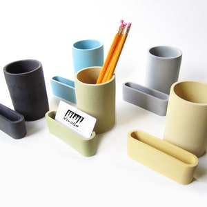 Op de afbeelding: Set van vijf betonnen bureau-organizers in verschillende kleuren, waaronder grijs, lichtblauw, geel en beige. De set bevat vier potlodenhouders en &eacute;&eacute;n visitekaartjeshouder. De potlodenhouders zijn cilindrisch en de visitekaartjeshouder is rechthoekig. De set is gerangschikt op een wit oppervlak.