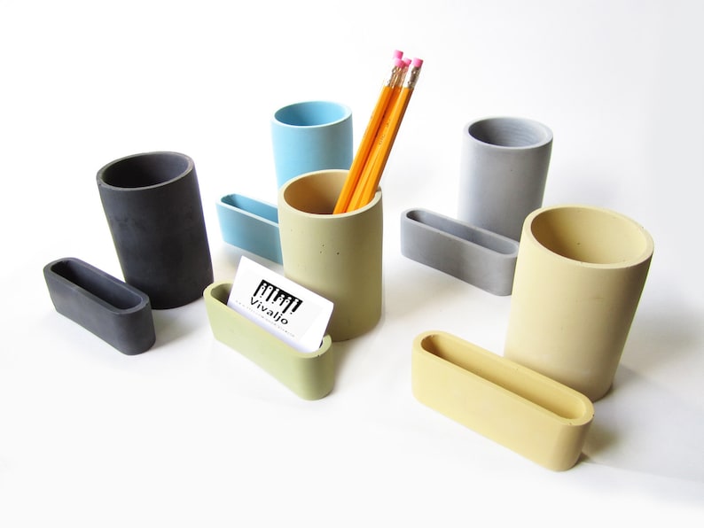 Op de afbeelding: Set van vijf betonnen bureau-organizers in verschillende kleuren, waaronder grijs, lichtblauw, geel en beige. De set bevat vier potlodenhouders en &eacute;&eacute;n visitekaartjeshouder. De potlodenhouders zijn cilindrisch en de visitekaartjeshouder is rechthoekig. De set is gerangschikt op een wit oppervlak.