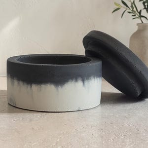May include: A two-tone, round concrete container with a lid. The container is dark gray at the top, fading into white at the bottom. The lid is dark gray and rests beside the container. The container is likely for storage or decorative use.