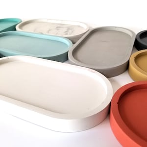 May include: A set of nine oval-shaped concrete trays in various colours, including white, grey, light blue, dark blue, yellow, and red. The trays are all the same size and shape, and they are arranged in a semi-circle.
