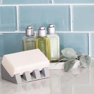 May include: A gray soap dish with five slots for bar soap. The dish is holding a white bar of soap. Three clear glass bottles with silver tops are in the background.