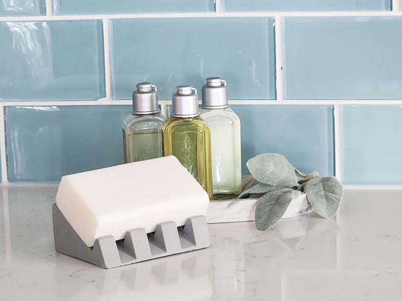 May include: A gray soap dish with five slots for bar soap. The dish is holding a white bar of soap. Three clear glass bottles with silver tops are in the background.