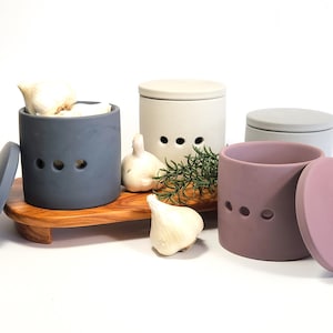 May include: Three ceramic garlic keepers with lids, one blue, one white and one purple. The blue keeper is filled with garlic cloves. The keepers have holes for air circulation. The keepers are on a wooden tray with a sprig of rosemary.
