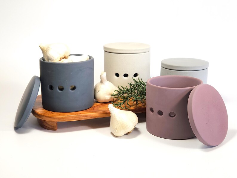 May include: Three ceramic garlic keepers with lids, one blue, one white and one purple. The blue keeper is filled with garlic cloves. The keepers have holes for air circulation. The keepers are on a wooden tray with a sprig of rosemary.