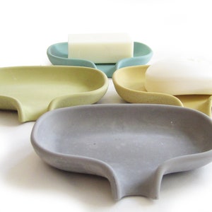 May include: Four soap dishes in different colors: light blue, yellow, gray, and beige. Each dish has a curved shape with a small drain in the bottom. Two of the dishes have bars of soap on top.