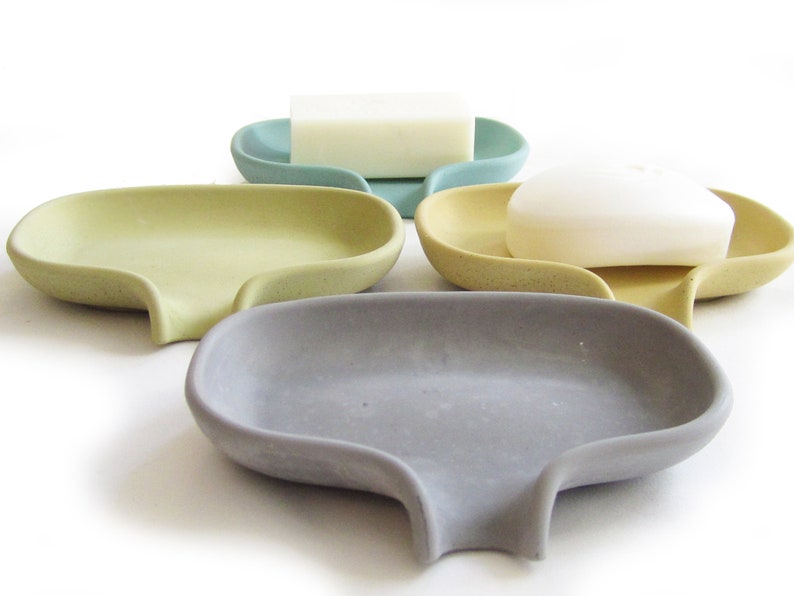 May include: Four soap dishes in different colors: light blue, yellow, gray, and beige. Each dish has a curved shape with a small drain in the bottom. Two of the dishes have bars of soap on top.