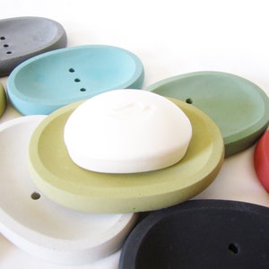 Puede incluir: Un conjunto de jaboneras ovaladas en varios colores, incluyendo gris, verde azulado, verde, amarillo, blanco, negro y rojo. Las jaboneras est&aacute;n hechas de hormig&oacute;n y tienen un acabado liso y redondeado. Una pastilla de jab&oacute;n blanca descansa sobre una de las jaboneras.