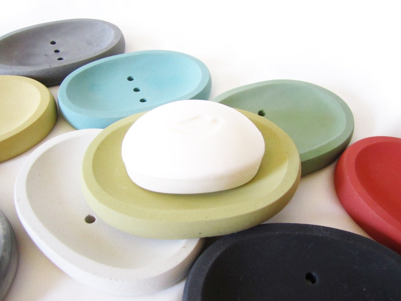 Puede incluir: Un conjunto de jaboneras ovaladas en varios colores, incluyendo gris, verde azulado, verde, amarillo, blanco, negro y rojo. Las jaboneras est&aacute;n hechas de hormig&oacute;n y tienen un acabado liso y redondeado. Una pastilla de jab&oacute;n blanca descansa sobre una de las jaboneras.