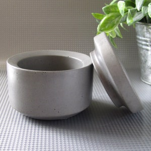 May include: A gray concrete container with a lid. The container is round and has a smooth, slightly textured surface. The lid is slightly domed and has a similar texture to the container.
