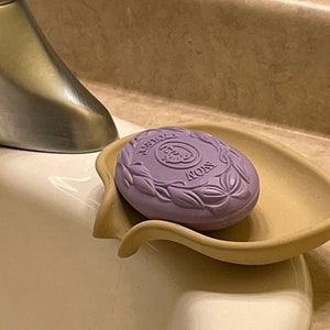 May include: A purple bar of soap with the word "Avon" embossed on it, sitting in a beige soap dish.