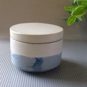 May include: A white ceramic jar with a lid, featuring a blue and white marbled design. The jar is sitting on a gray textured surface.