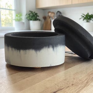 May include: A two-tone, black and white concrete container with a matching lid. The container is cylindrical with a dip-dye effect, transitioning from black to white. The lid is a flat, oval shape with a ridged design. The container sits on a wooden surface.