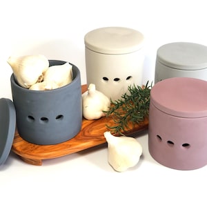 May include: Three ceramic garlic keepers in different colours: grey, light grey, and pink. Each keeper has a lid and three air holes. The grey keeper is filled with garlic cloves. The keepers are on a wooden tray with a sprig of rosemary.