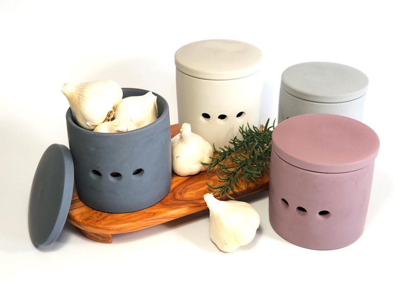 May include: Three ceramic garlic keepers in different colours: grey, light grey, and pink. Each keeper has a lid and three air holes. The grey keeper is filled with garlic cloves. The keepers are on a wooden tray with a sprig of rosemary.