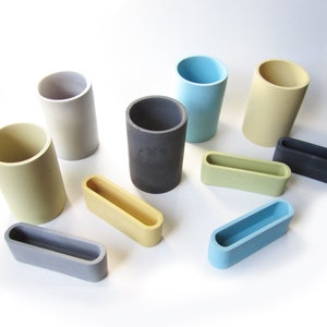 Op de afbeelding: Een set van tien kleine, cilindrische en rechthoekige containers in verschillende kleuren, waaronder grijs, lichtblauw, geel en donkergrijs. De containers zijn gemaakt van beton en hebben een gladde, matte afwerking.
