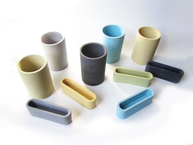 Op de afbeelding: Een set van tien kleine, cilindrische en rechthoekige containers in verschillende kleuren, waaronder grijs, lichtblauw, geel en donkergrijs. De containers zijn gemaakt van beton en hebben een gladde, matte afwerking.
