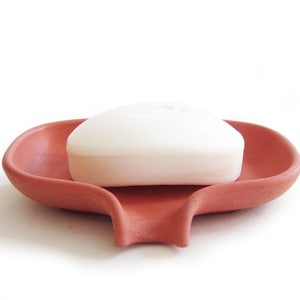 May include: A terracotta soap dish with a white bar of soap. The dish has a curved design and a drainage spout.