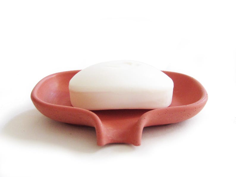 May include: A terracotta soap dish with a white bar of soap. The dish has a curved design and a drainage spout.