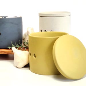 May include: Three ceramic garlic keepers with lids. One is blue, one is white, and one is yellow. The yellow keeper is open, revealing the lid.