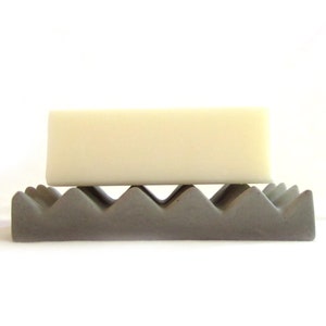 May include: A gray concrete soap dish with a wavy design, holding a white bar of soap.