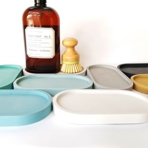 May include: A set of seven oval-shaped concrete trays in various colours, including grey, blue, yellow, red, and black. The trays are arranged in a semi-circle on a white surface.