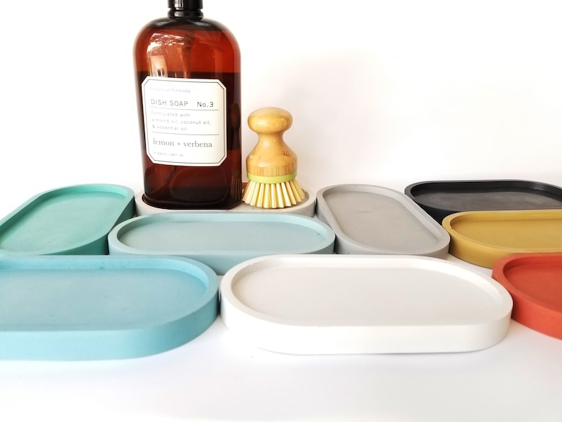 May include: A set of seven oval-shaped concrete trays in various colours, including grey, blue, yellow, red, and black. The trays are arranged in a semi-circle on a white surface.