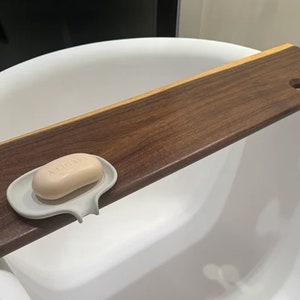 May include: A wooden bath tray with a soap dish and a hole for a glass. The tray is made of dark wood and has a light wood accent. The soap dish is white and has a soap bar inside.
